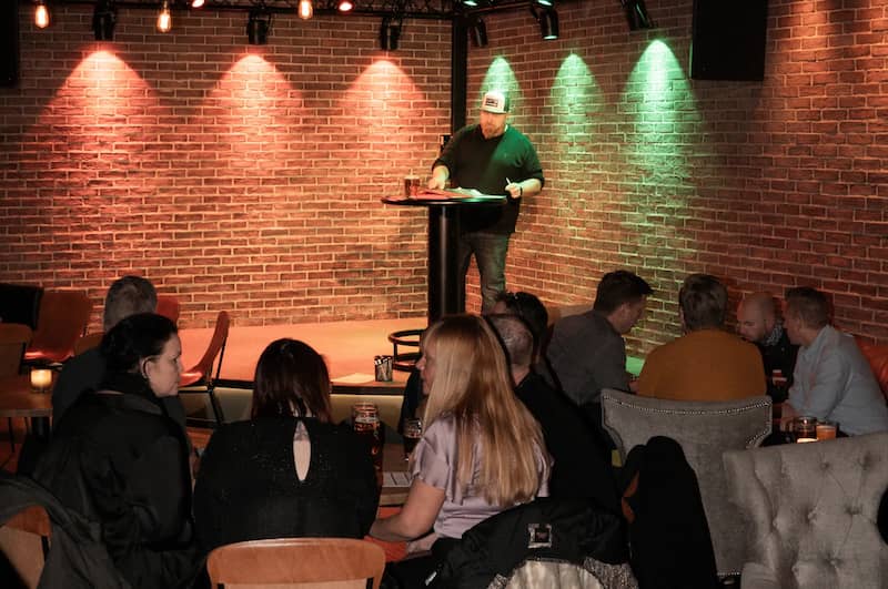 En man i keps står på en scen i strålkastare vid ett ståbord, inför publik