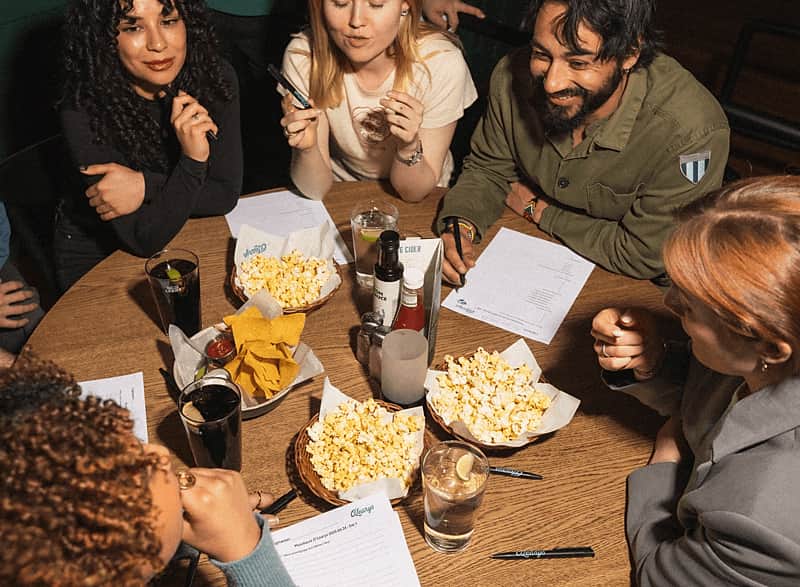 Sällskap med vänner som sitter med papper och penna. Har popcorn, nachos och dryck på bordet